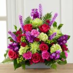 A lush and vibrant "Warm Embrace" floral arrangement featuring a mix of magenta roses, purple liatris, lime green spider mums, and pink carnations in a low gray rectangular container.