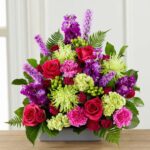 A vibrant and lush floral arrangement titled "Warm Embrace," featuring a mix of hot pink roses, purple stock flowers, lime green spider mums, and pink carnations in a sleek gray rectangular container.