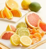 A vibrant assortment of sliced and whole citrus fruits, including oranges, lemons, limes, pink grapefruits, and mandarin segments, arranged on white plates over a light-colored wooden surface.