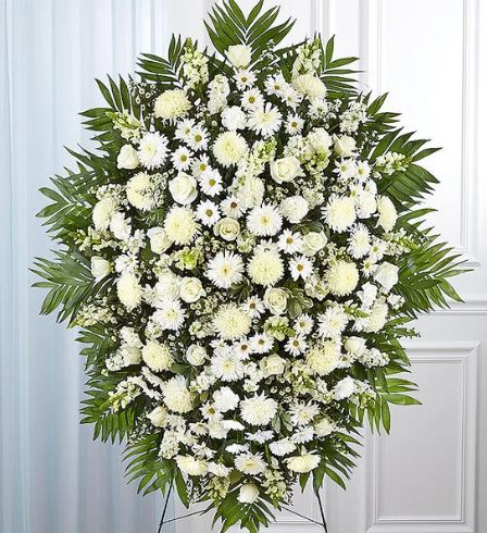 A large, elegant white funeral standing spray arrangement featuring roses, lilies, chrysanthemums, and snapdragons set against lush green palm fronds on a wire easel.