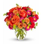 A vibrant bouquet of orange roses, hot pink gerbera daisies, red carnations, and small yellow accent flowers arranged in a clear glass vase with visible green stems.