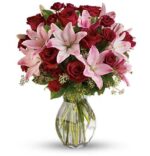 Elegant floral arrangement in a clear glass vase featuring deep red roses, light pink lilies, pink alstroemeria, and green filler leaves