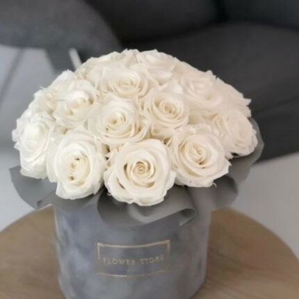 A luxurious arrangement of pristine white roses tightly packed in a round, grey velvet hatbox with a gold-embossed label.