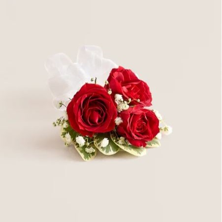 Red spray rose corsage with three deep red roses, white baby’s breath, variegated greenery, and white satin ribbon on a cream background.