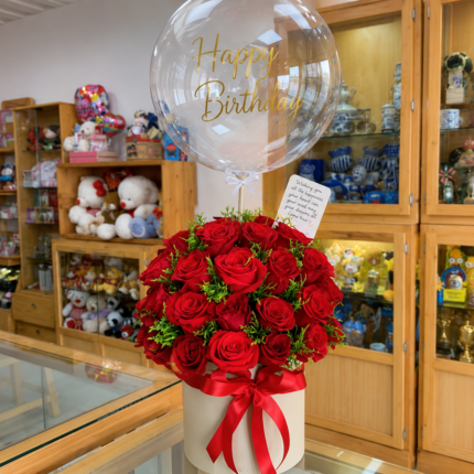 A luxurious floral gift arrangement featuring a large bouquet of red roses in a cylindrical grey box, topped with a clear bubble balloon filled with silver confetti.