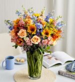 A vibrant and large spring flower bouquet in a tall glass vase, featuring peach roses, yellow sunflowers, blue delphiniums, and pink alstroemeria, set on a white table next to a blue mug and a book.