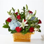 A vibrant floral arrangement featuring red roses, white lilies, and light blue hydrangeas set in a woven light-brown basket against a white background.