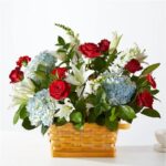 A vibrant floral arrangement featuring red roses, white lilies, and light blue hydrangeas set in a rectangular woven wicker basket.