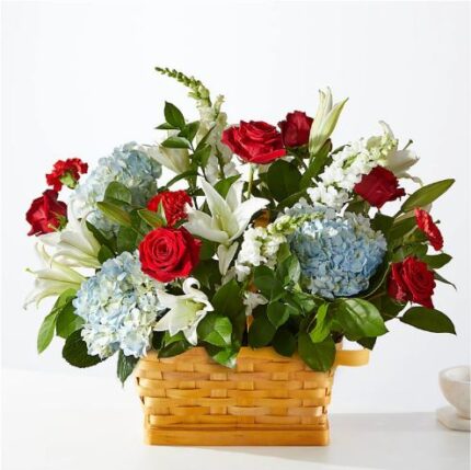 A vibrant floral arrangement featuring red roses, white lilies, and light blue hydrangeas set in a rectangular woven wicker basket.