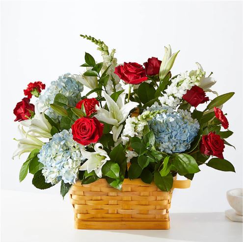 A vibrant floral arrangement featuring red roses, white lilies, and light blue hydrangeas set in a rectangular woven wicker basket.