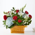 A lush floral arrangement featuring red roses, light blue hydrangeas, white lilies, and snapdragons in a light wood woven basket.
