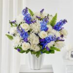 Large floral arrangement featuring white roses, white chrysanthemums, and vibrant blue delphiniums in a white fluted ceramic vase