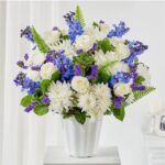 A lush floral arrangement of white roses, white chrysanthemums, and blue delphiniums in a white fluted ceramic vase.