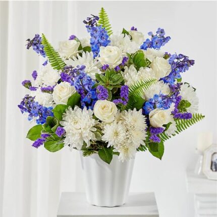 A lush floral arrangement of white roses, white chrysanthemums, and blue delphiniums in a white fluted ceramic vase.