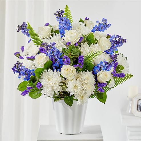 A lush floral arrangement of white roses, white chrysanthemums, and blue delphiniums in a white fluted ceramic vase.