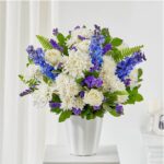 Elegant floral bouquet featuring white roses, chrysanthemums, and blue delphiniums in a white ceramic vase, suitable for funeral or sympathy services.