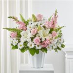 A lush floral arrangement featuring light pink roses, white lilies, pink snapdragons, and white daisies in a fluted white ceramic vase, set against a neutral background.