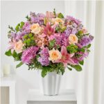 A large, lush floral arrangement in a white fluted ceramic vase, featuring pink lilies, peach roses, and purple chrysanthemums.