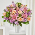 A large, vibrant bouquet of pink lilies, peach roses, and purple flowers arranged in a white fluted ceramic vase.
