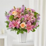 A lush floral arrangement in a white fluted vase featuring pink lilies, peach roses, and purple chrysanthemums.