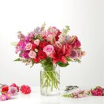 A vibrant bouquet of pink, red, and purple flowers including roses and lilies in a clear cylindrical glass vase on a white surface.