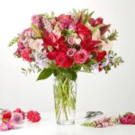 A vibrant and lush bouquet of mixed flowers, featuring red lilies, pink roses, and purple snapdragons, arranged in a tall clear glass vase on a white surface.
