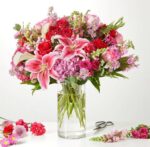A vibrant and lush floral arrangement in a clear cylindrical glass vase featuring pink lilies, red roses, and purple hydrangeas.
