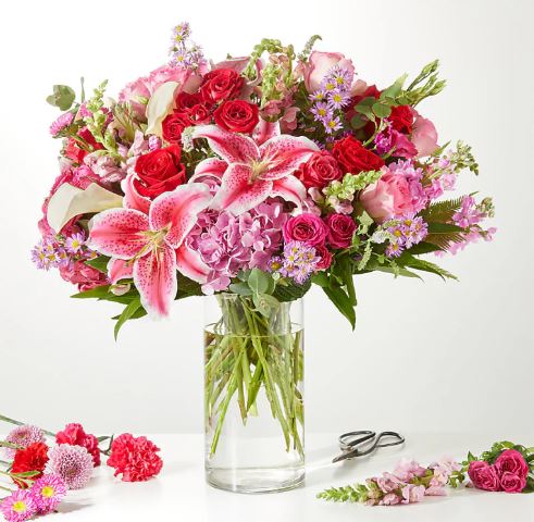 A vibrant and lush floral arrangement in a clear cylindrical glass vase featuring pink lilies, red roses, and purple hydrangeas.