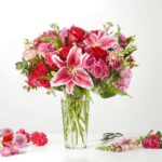 A large, vibrant bouquet of pink and red flowers, including stargazer lilies, roses, and carnations, arranged in a clear glass vase on a white surface.