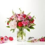 A vibrant floral arrangement featuring pink hydrangeas, red carnations, and purple roses in a clear cylindrical glass vase on a white surface.