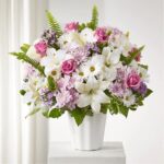 A lush floral arrangement in a white fluted vase featuring white lilies, pink roses, white daisies, and purple chrysanthemums accented with green ferns.