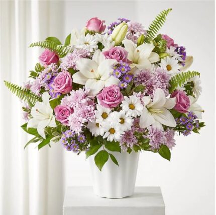 A large, lush floral arrangement featuring white lilies, pink roses, purple chrysanthemums, and white daisies in a fluted white ceramic vase, set against a soft white background.