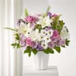 A lush floral arrangement of white lilies, pink roses, and purple daisies in a white vase.